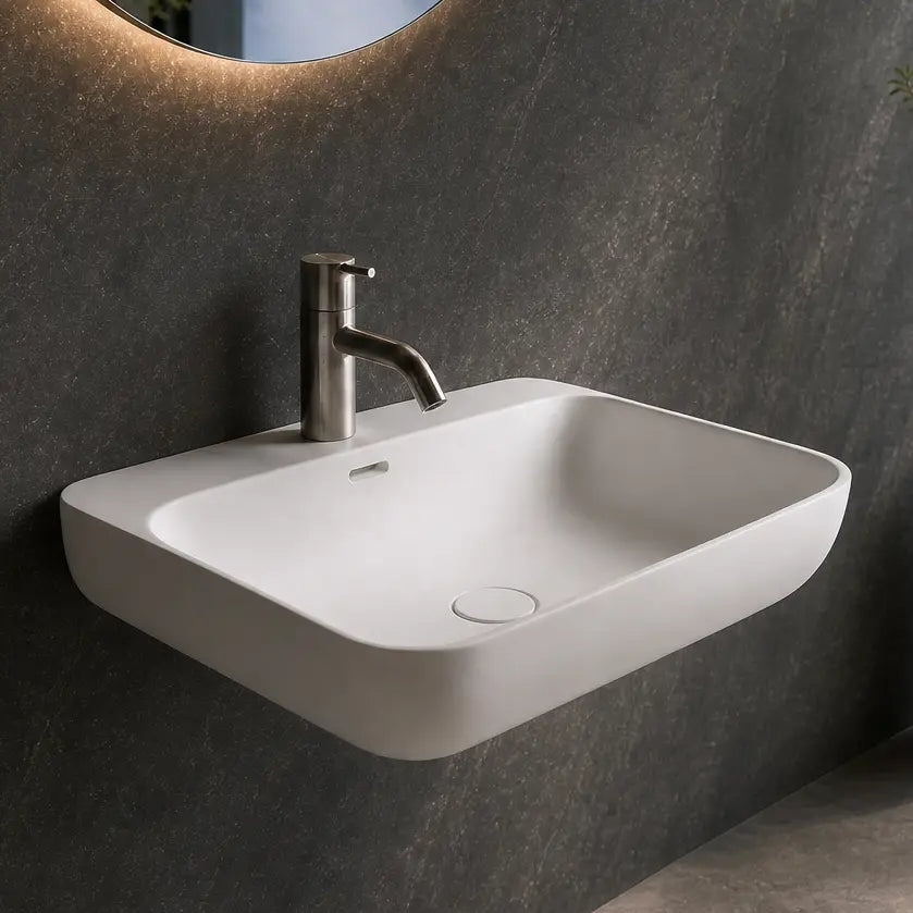 A modern wall-mounted white sink with a chrome faucet in a dark gray bathroom, featuring a large oval mirror with warm lighting and a large window overlooking a waterfall landscape.