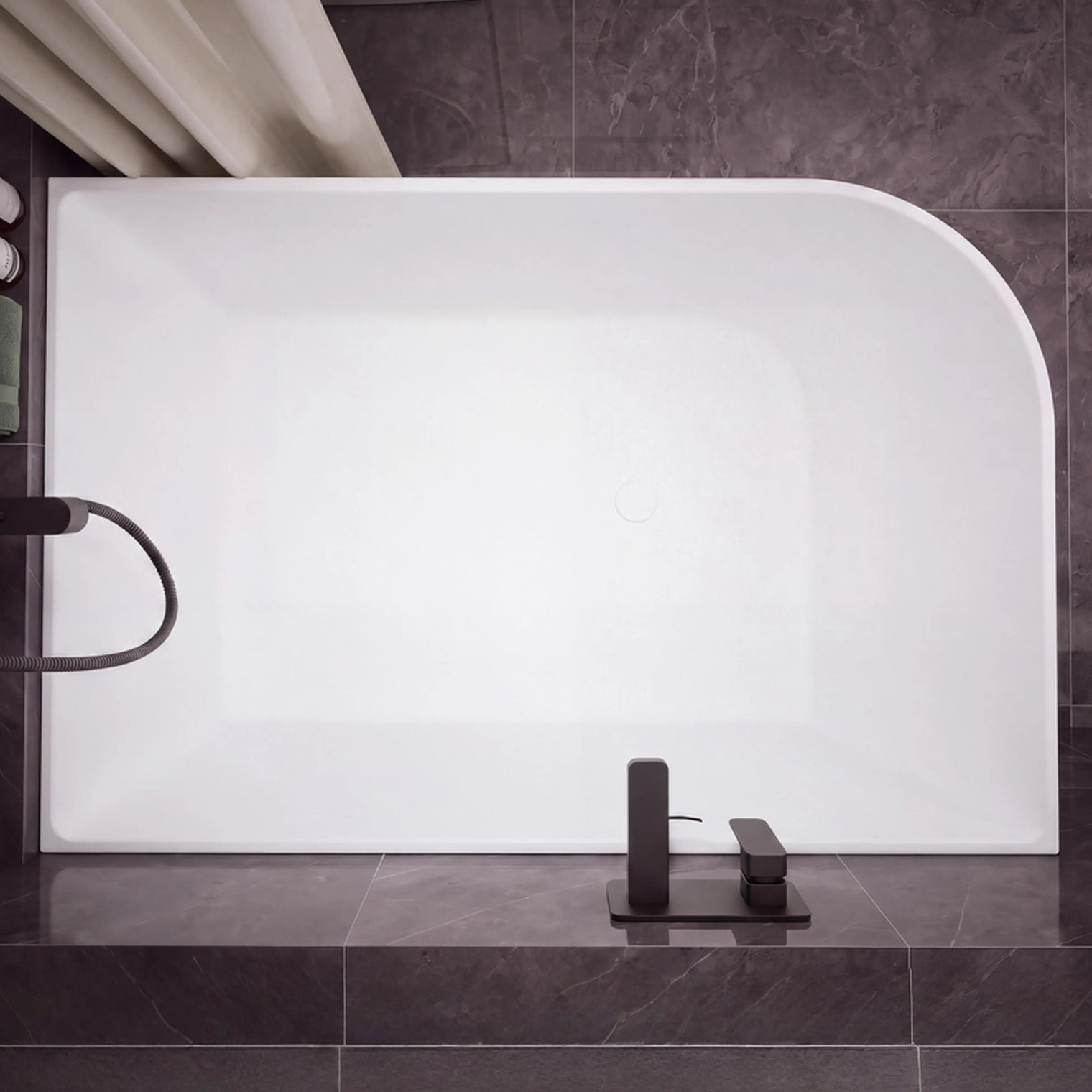 A modern freestanding rectangular bathtub with rounded corners, shown installed against dark gray tiled walls and floor. The tub is matte white, with matte black fixtures mounted on the right side and a matching black faucet centered at the bottom.