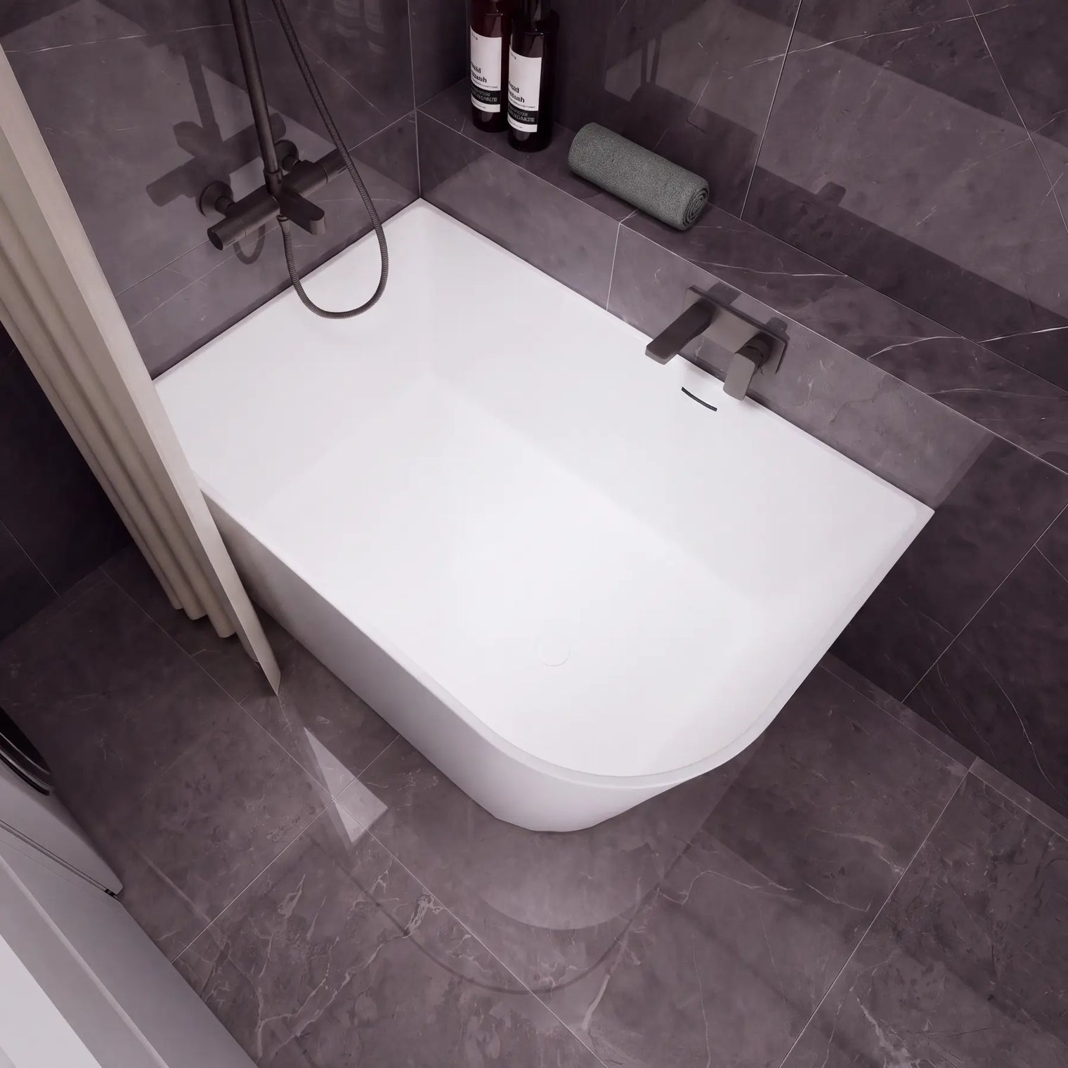A modern white freestanding bathtub with a dark gray tiled bathroom setting, featuring a wall-mounted chrome faucet, a handheld shower attachment, and a beige curtain on the left.