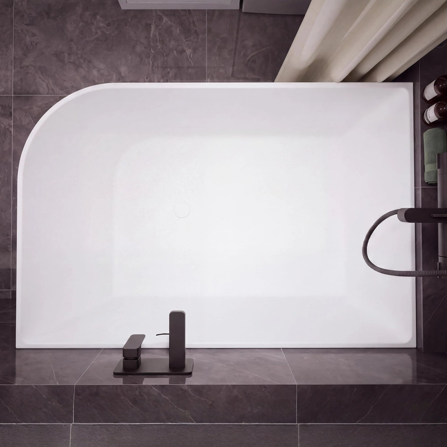 A modern freestanding rectangular bathtub with rounded corners, shown installed against dark gray tiled walls and floor. The tub is matte white, with matte black fixtures mounted on the right side and a matching black faucet centered at the bottom.