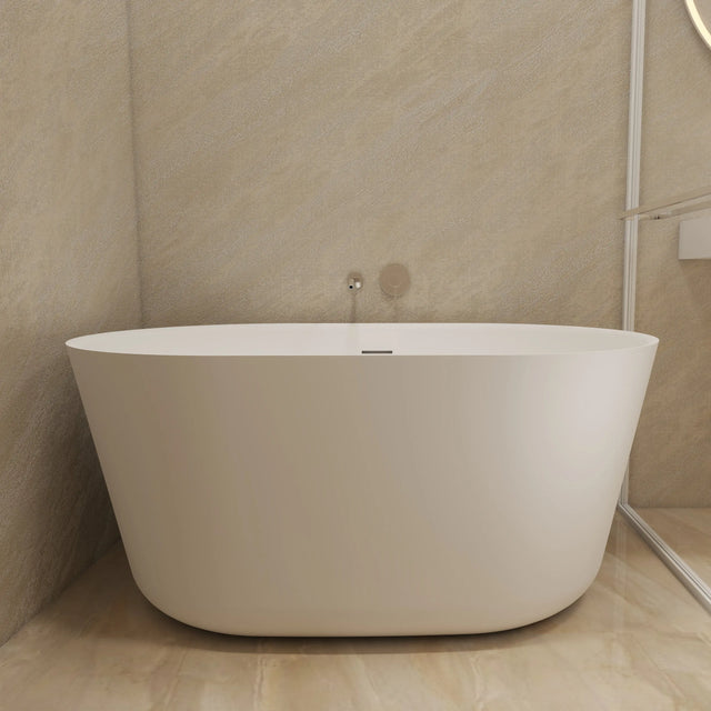 A modern freestanding oval bathtub in matte white, set in a minimalist bathroom with beige stone walls and floor, next to a glass shower screen.