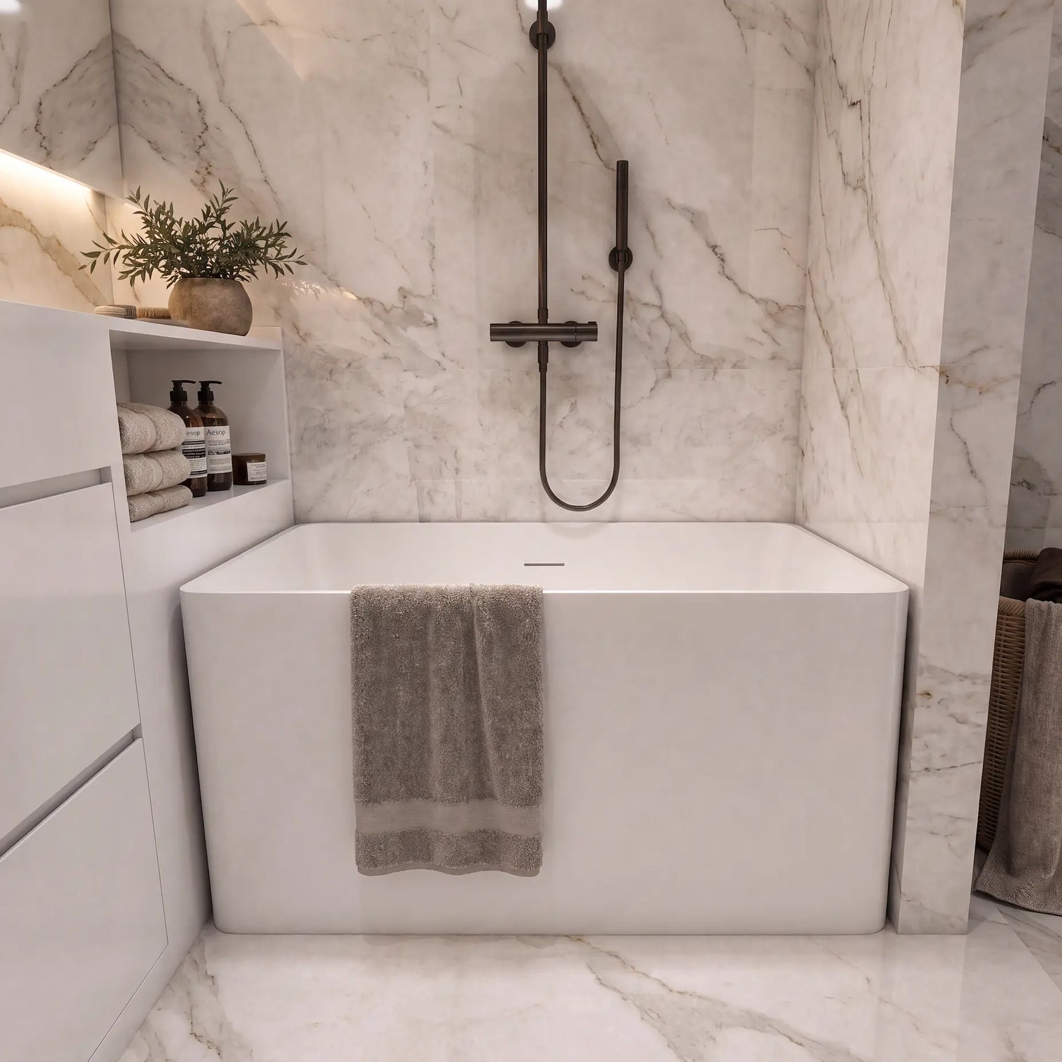 A modern freestanding white bathtub in a luxury bathroom with white marble walls and floor, featuring a matte black handheld shower, a folded taupe towel draped over the tub edge, and a matching towel basket on the right.