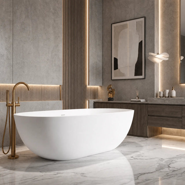 A modern freestanding oval bathtub in white with a brushed gold faucet, set in a contemporary bathroom with gray stone walls and marble flooring.