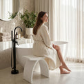 Woman in a white robe sitting by a bathtub in a modern bathroom.