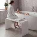 Two women in a modern bathroom with one sitting on a bench by a bathtub and the other lying inside.