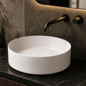 White round sink on a dark marble countertop with a mirror and bottles in the background.