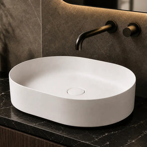 White oval sink on a dark marble countertop with a modern mirror and decorative bottles.