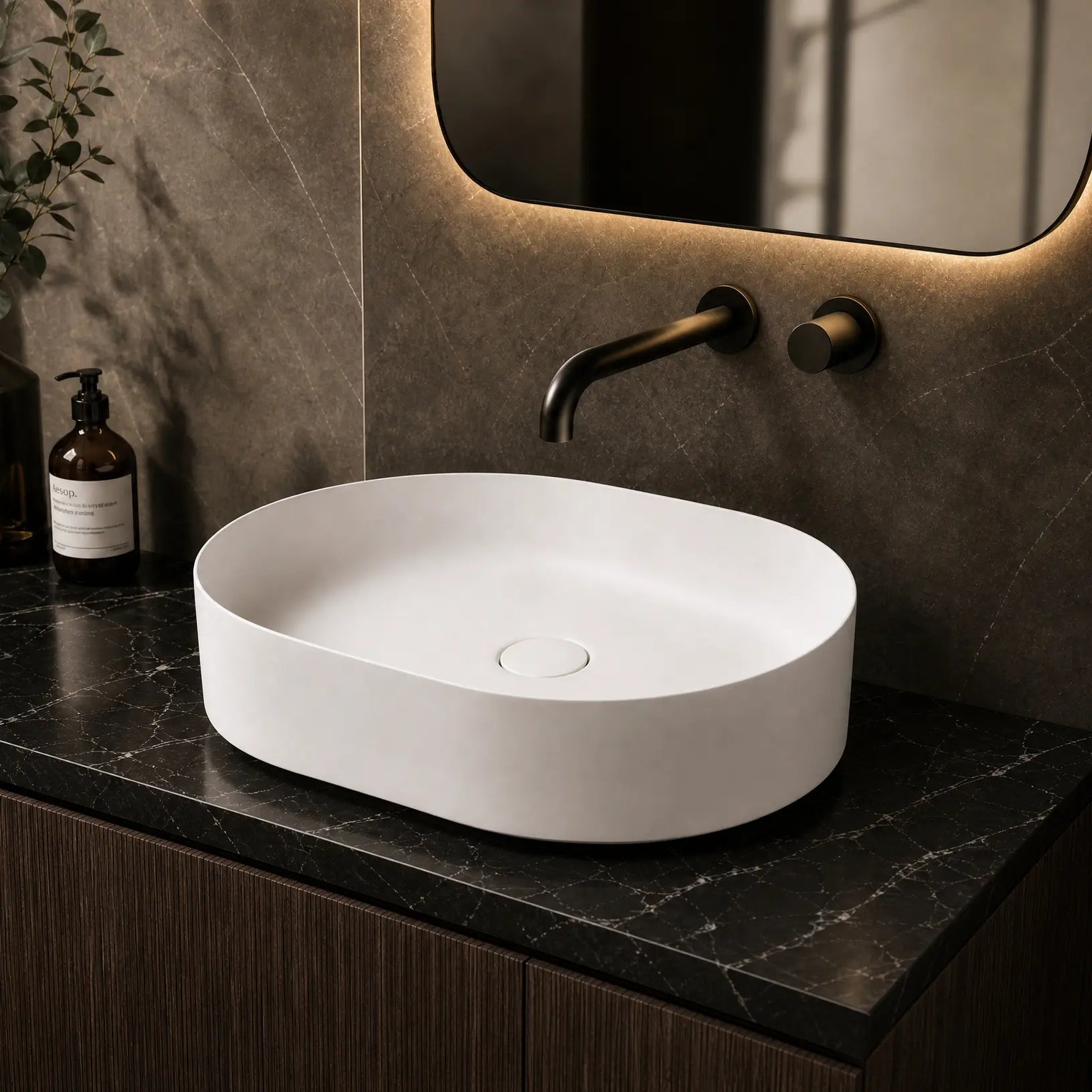 White oval sink on a dark marble countertop with a modern mirror and decorative bottles.