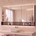 A modern bathroom vanity featuring a large rectangular wall mirror with a dark reflective base, mounted above a floating stone sink and wooden console. The room has warm beige textured walls, built-in shelves with candles and decor, and a window on the left with sheer curtains.