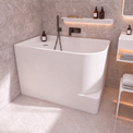 A modern freestanding bathtub in a light gray bathroom with matching tiled walls and floor, featuring a matte black wall-mounted faucet and small bottles on a shelf.