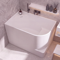 A modern freestanding bathtub in a minimalist bathroom setting with gray marble-look tiled walls and floor, a white vanity, and a small white stool.