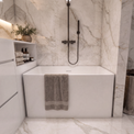 A modern freestanding white bathtub in a luxury bathroom with white marble walls and floor, featuring a matte black handheld shower, a folded taupe towel draped over the tub edge, and a matching towel basket on the right.