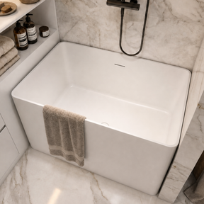 A modern freestanding white bathtub installed in a marble bathroom, with a black wall-mounted shower fixture, beige towels, a vanity shelf with toiletries, and a woven basket on the floor.