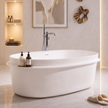 A modern freestanding oval bathtub in a bright, minimalist bathroom with warm ambient lighting, a wall-mounted sink, a round backlit mirror, framed wall art, and a small woven basket with a towel.