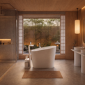 A modern freestanding oval bathtub in a minimalist, warm-toned bathroom with wood walls, dual pedestal sinks, a glass shower enclosure, and a large window overlooking bamboo.