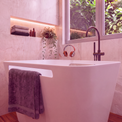 A modern freestanding bathtub in a pink-toned bathroom, with a dark towel draped over the side, a wall-mounted shelf with folded towels and bottles, a window with greenery outside, and a dark faucet. A pair of orange headphones rests on the tub edge.