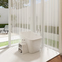 A modern freestanding white bathtub placed in a bright, minimalist bathroom with sheer white curtains, large floor-to-ceiling windows, and an outdoor patio visible beyond.