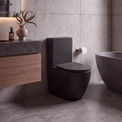 A modern bathroom interior featuring a matte black wall-mounted toilet, a dark floating vanity with wood grain, gray stone-look tiles, a freestanding oval bathtub, and brass fixtures.