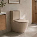 A modern 2-piece ceramic toilet in a warm beige or taupe color, shown installed in a minimalist bathroom setting with a wood-look tiled shower area and neutral-toned walls.