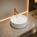 White round Solid Surfaces sink with gold faucet on a beige countertop in a bathroom setting.