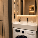A modern bathroom vanity setup featuring a white Samsung washing machine integrated beneath a white sink with a chrome faucet, set against warm beige stone walls with built-in shelving and a glass shower enclosure.