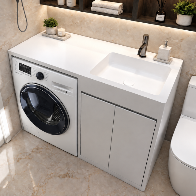 A modern white bathroom vanity cabinet with an integrated sink, next to a washing machine, in a marble-look bathroom. The vanity is styled with a soap dispenser, a small plant, and a hair dryer is mounted on the tiled wall.