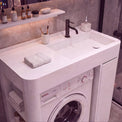 A white integrated washing and sink vanity unit in a modern bathroom, featuring a Bosch front-loading washing machine beneath a double basin sink with a matte black faucet. The unit is styled with folded towels, toiletries on a shelf, and a glass shower enclosure in the background.