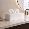 A white rectangular tissue box cover holding white tissue paper, placed on a light marble bathroom countertop near a sink, faucet, and round mirror.