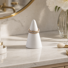 A white cone-shaped ring display stand holding a rose gold ring with clear stones, set on a white marble countertop with gold and white bathroom accessories in the background.