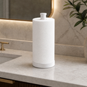 A white cylindrical countertop paper towel holder with a textured paper towel roll inserted, shown on a light marble bathroom counter beside a sink and a potted plant.