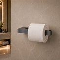 Black toilet paper holder with a roll of toilet paper on a beige tiled bathroom wall.
